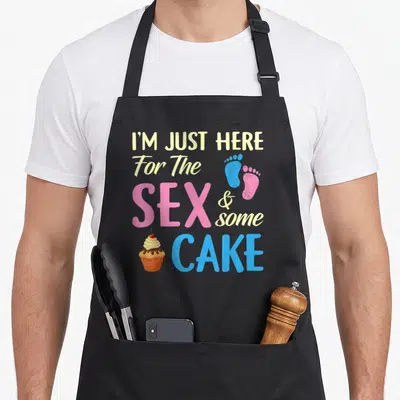 Gender cake apron - Stacked text I'M JUST HERE For The SEX & some CAKE with cupcake and blue pink footprints — shown on a black cotton canvas apron against a white background