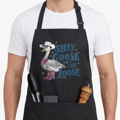 Cute goose silly apron - Cartoon goose in cowboy hat and pink boots next to blue text SILLY GOOSE ON THE LOOSE — shown on a black cotton canvas apron against a white background