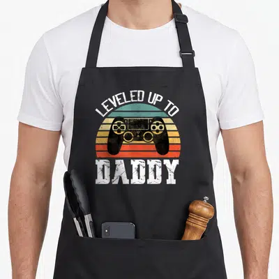Leveled father day apron - Retro striped sun with black game controller and LEVELED UP TO DADDY text — shown on a black cotton canvas apron against a white background