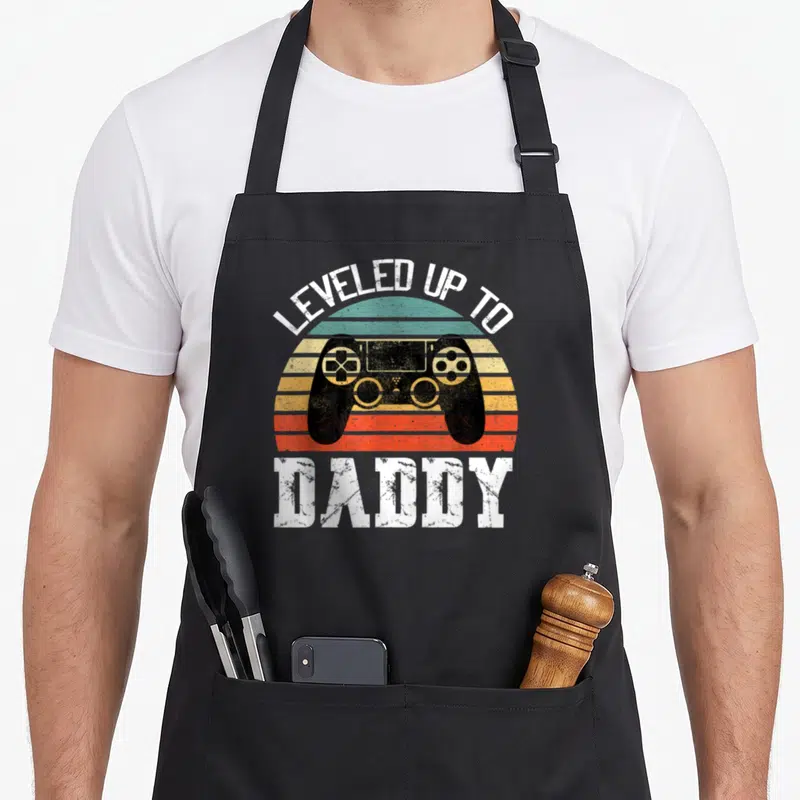 Leveled father day apron - Retro striped sun with black game controller and LEVELED UP TO DADDY text — shown on a black cotton canvas apron against a white background