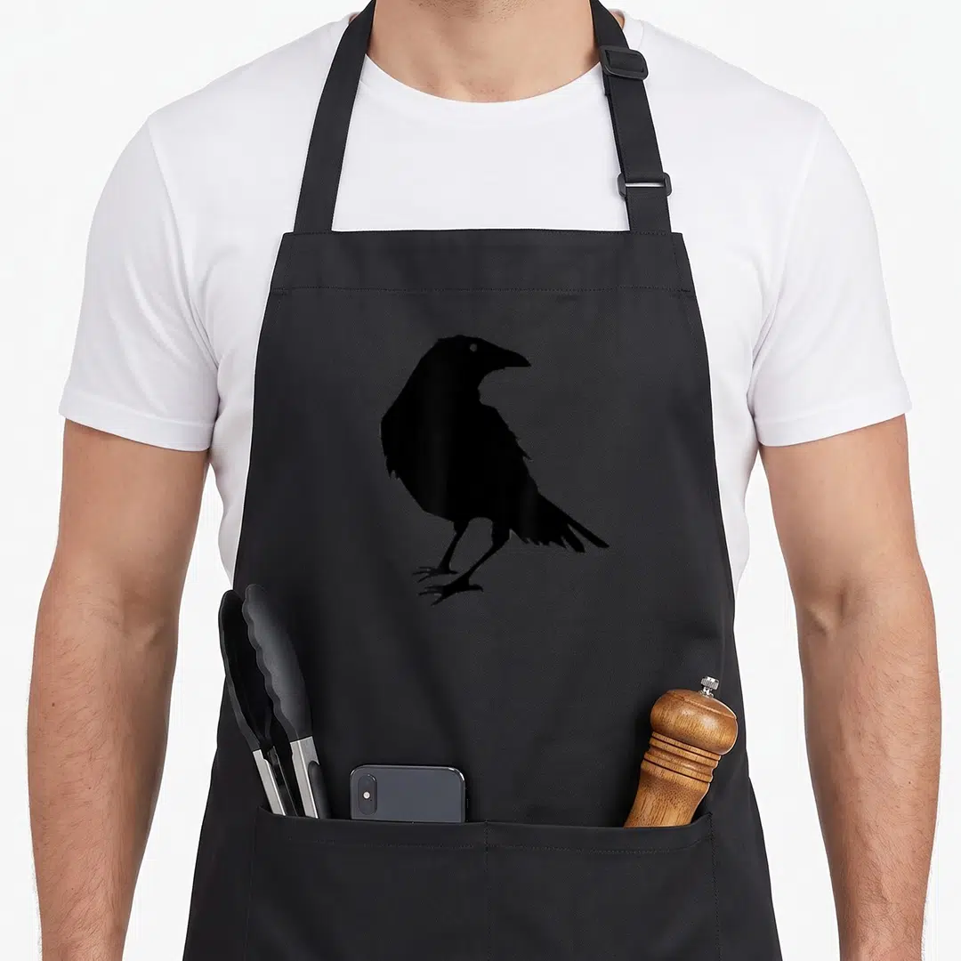 Beautiful bird aprons - Minimalist black crow silhouette standing in profile with visible eye and talons — shown on a black cotton canvas apron against a white background