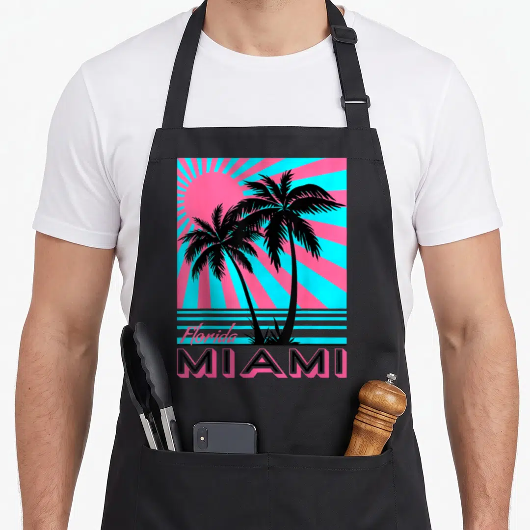Miami tree apron - Two black palm trees against pink sun with cyan rays and Florida Miami text — shown on a black cotton canvas apron against a white background