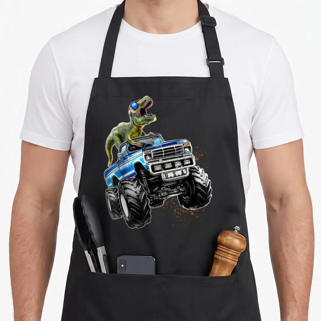 Patriotic truck apron - Green T-Rex in sunglasses on blue monster truck with USA bumper text and brown mud splatters — shown on a black cotton canvas apron against a white background