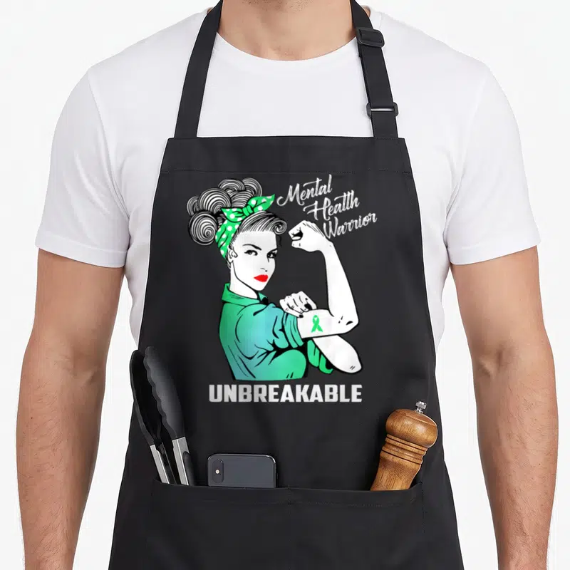 Warrior mental health awareness apron - Cartoon woman flexing arm with green ribbon and Mental Health Warrior — shown on a black cotton canvas apron against a white background