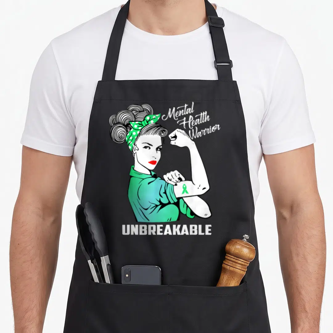 Warrior mental health awareness apron - Cartoon woman flexing arm with green ribbon and Mental Health Warrior — shown on a black cotton canvas apron against a white background