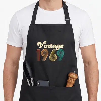 50th birthday party apron - Vintage 1969 text in beige, orange, yellow, and blue retro font stacked vertically — shown on a black cotton canvas apron against a white background