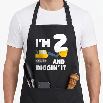 Excavator truck apron - Cartoon yellow excavator with I'M 2 AND DIGGIN' IT text in white — shown on a black cotton canvas apron against a white background