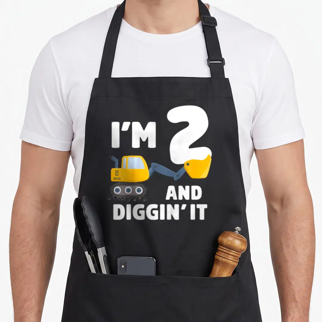 Excavator truck apron - Cartoon yellow excavator with I'M 2 AND DIGGIN' IT text in white — shown on a black cotton canvas apron against a white background