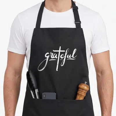 Grateful cross apron - White script text grateful with t shaped like a cross — shown on a black cotton canvas apron against a white background