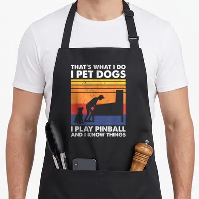Pinball dog apron - Retro striped sunset with grey person dog silhouette and white text about pinball and dogs — shown on a black cotton canvas apron against a white background