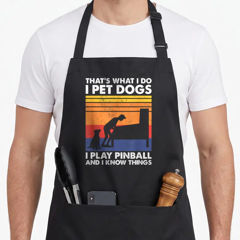 Pinball dog apron - Retro striped sunset with grey person dog silhouette and white text about pinball and dogs — shown on a black cotton canvas apron against a white background