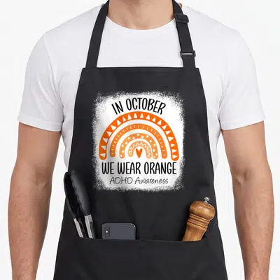 October rainbow apron - Orange rainbow with white patterns and black text In October We Wear Orange ADHD Awareness — shown on a black cotton canvas apron against a white background