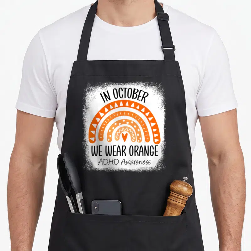 October rainbow apron - Orange rainbow with white patterns and black text In October We Wear Orange ADHD Awareness — shown on a black cotton canvas apron against a white background