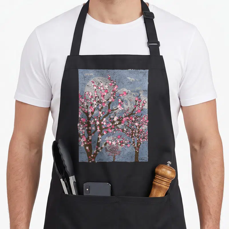 Japan cherry print apron - Watercolor cherry blossom trees with pink flowers, brown branches, and a large grey moon — shown on a black cotton canvas apron against a white background