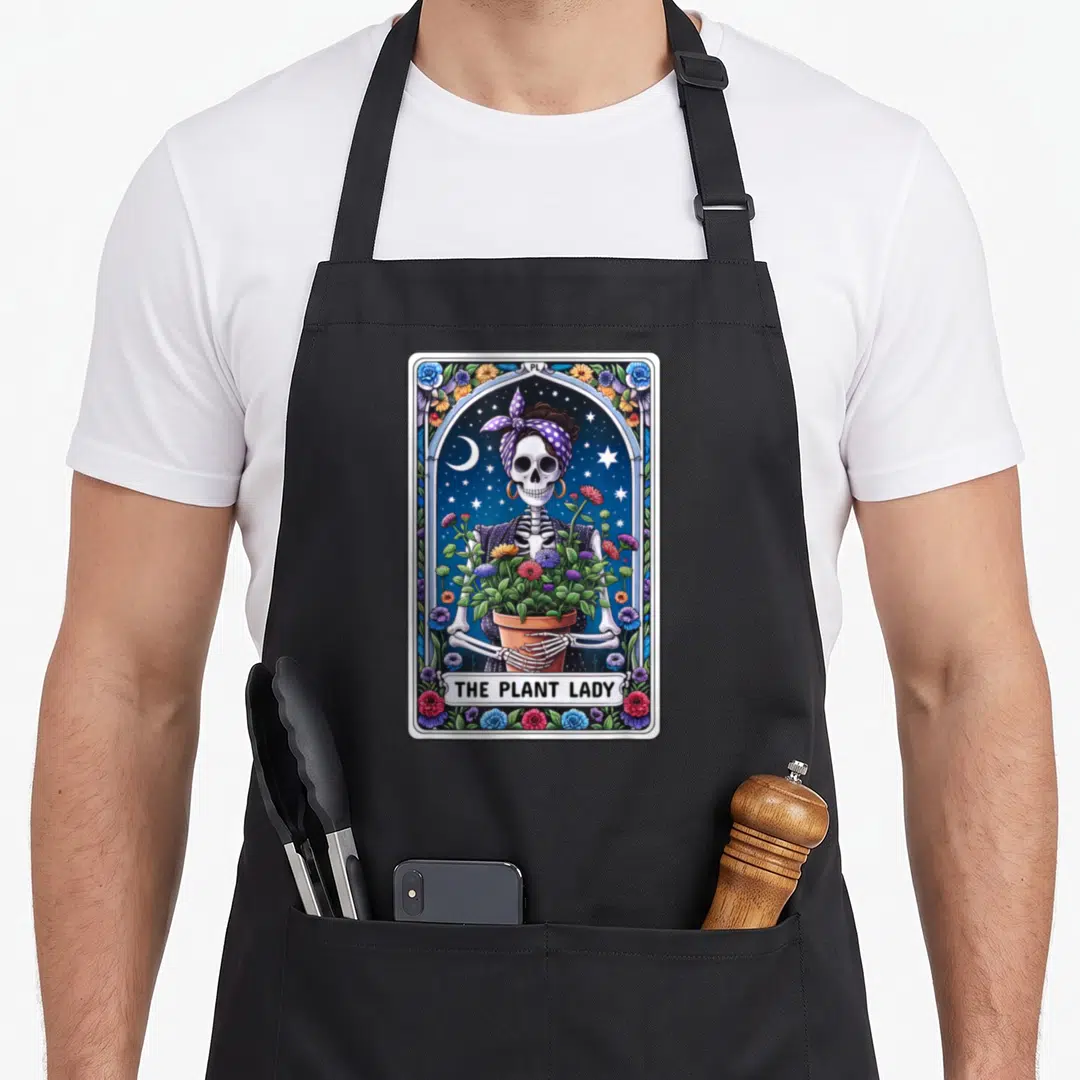Tarot plant lady apron - Skeleton woman in purple bandana holding potted flowers with THE PLANT LADY text in decorative — shown on a black cotton canvas apron against a white background