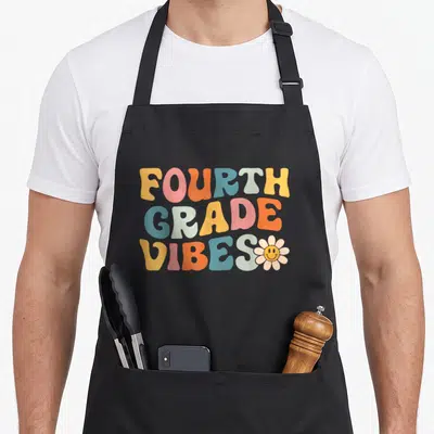 Fourth grade vibes apron - Retro bubbly text reading Fourth Grade Vibes with smiley flower in yellow pink orange blue — shown on a black cotton canvas apron against a white background