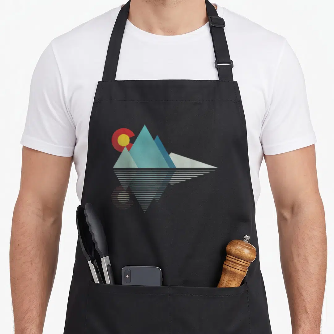 Flag colorado apron - Three blue mountain peaks with red and yellow sun and horizontal line reflection — shown on a black cotton canvas apron against a white background