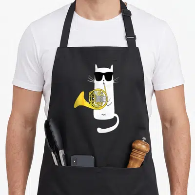 French cute cat apron - White cartoon cat wearing black sunglasses playing a yellow french horn — shown on a black cotton canvas apron against a white background