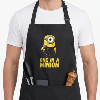 Despicable portrait general apron - Yellow one-eyed minion character standing above yellow ONE IN A MINION text — shown on a black cotton canvas apron against a white background