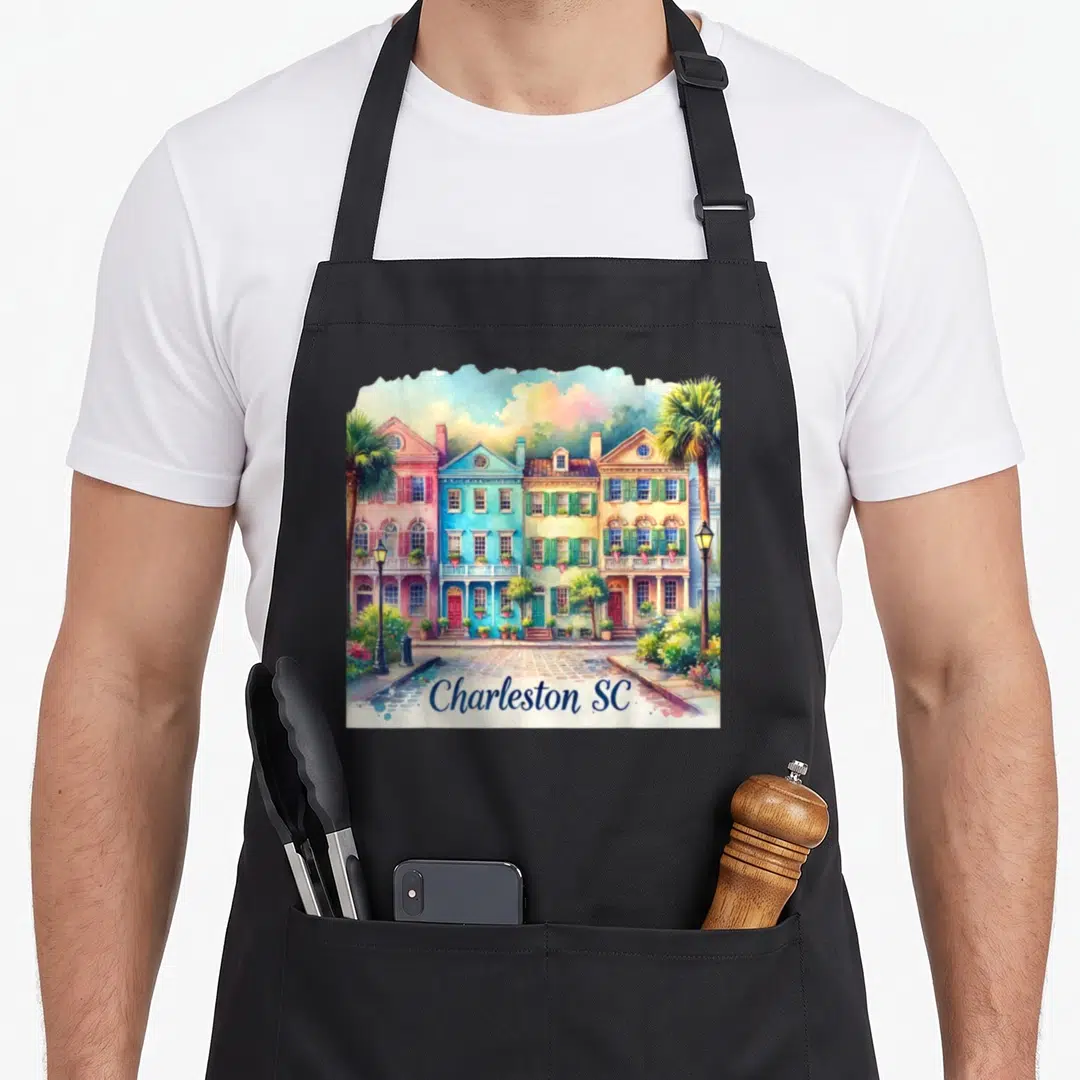 Charleston colorful aprons - Watercolor row of pink, blue, yellow, and green houses above a street with Charleston SC — shown on a black cotton canvas apron against a white background