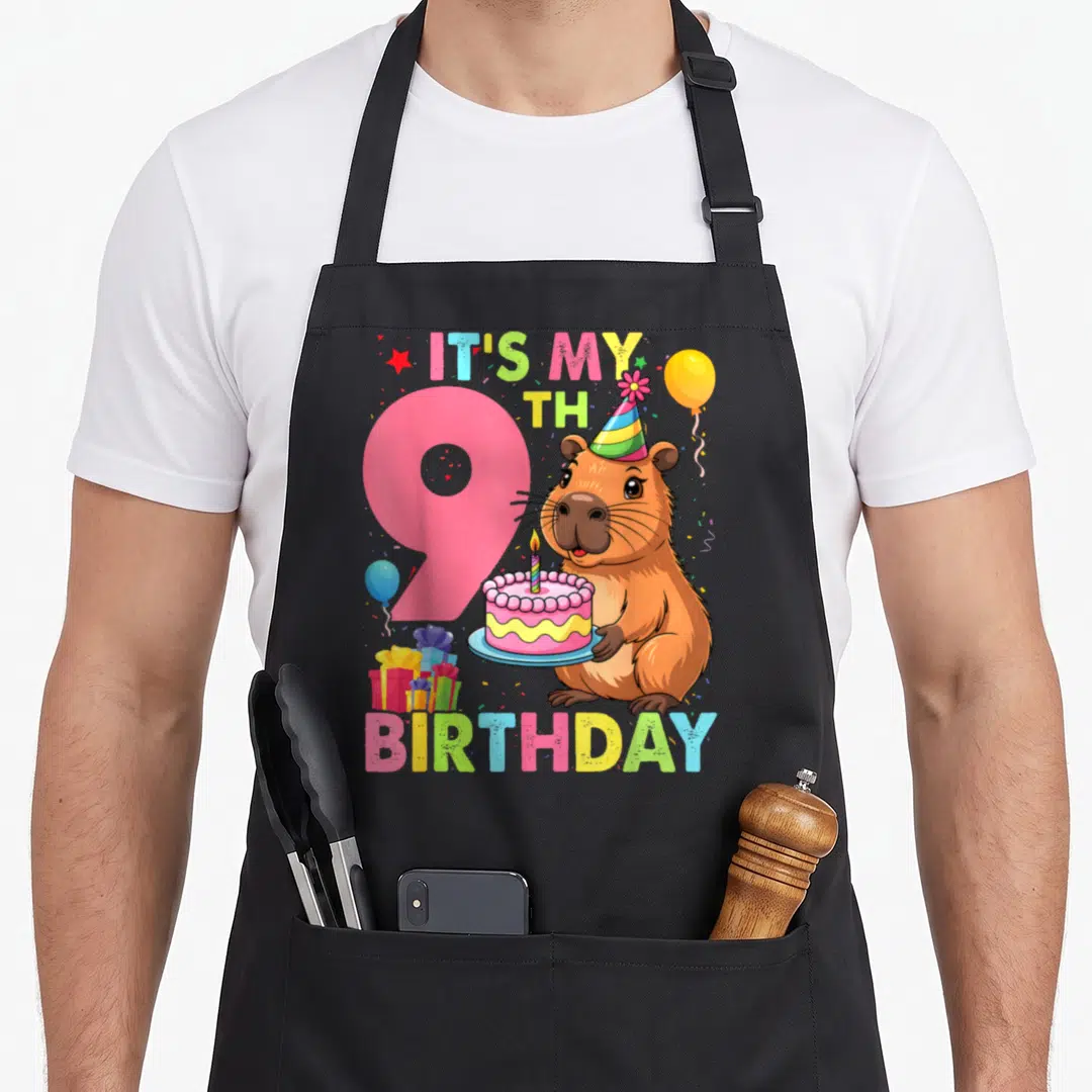 9th capybara birthday apron - Cartoon capybara holding cake next to pink number 9 with balloons and gifts — shown on a black cotton canvas apron against a white background