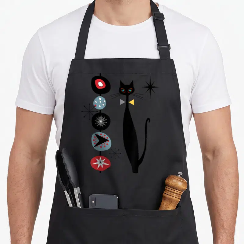 Era modern apron - Tall black cat next to five stacked abstract shapes and three starbursts in retro colors — shown on a black cotton canvas apron against a white background