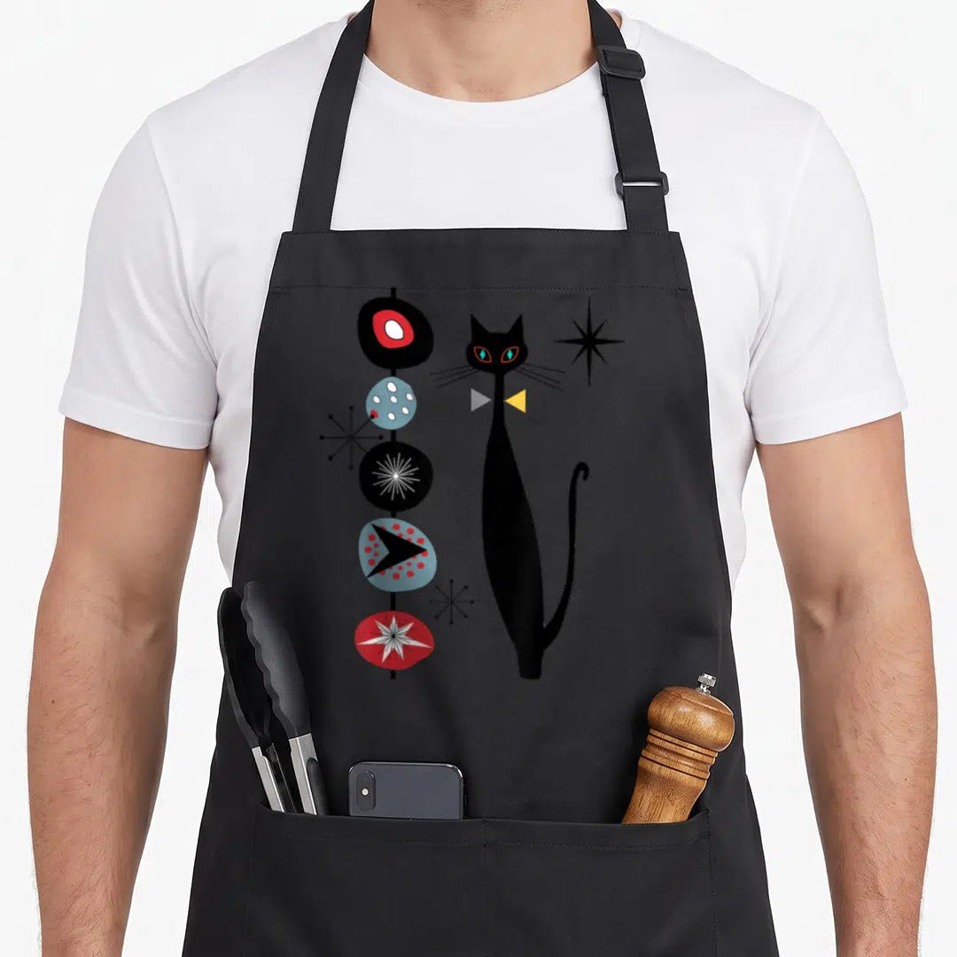 Era modern apron - Tall black cat next to five stacked abstract shapes and three starbursts in retro colors — shown on a black cotton canvas apron against a white background