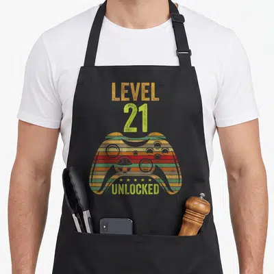 Cool level birthday apron - Striped game controller with LEVEL 21 UNLOCKED text and five stars in distressed orange red — shown on a black cotton canvas apron against a white background