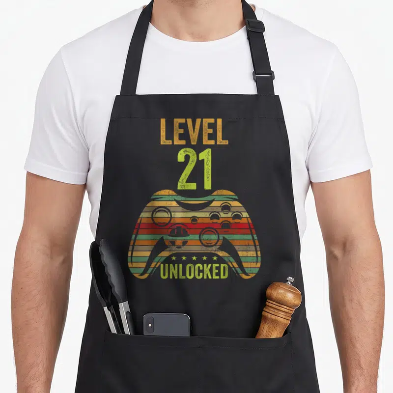 Cool level birthday apron - Striped game controller with LEVEL 21 UNLOCKED text and five stars in distressed orange red — shown on a black cotton canvas apron against a white background