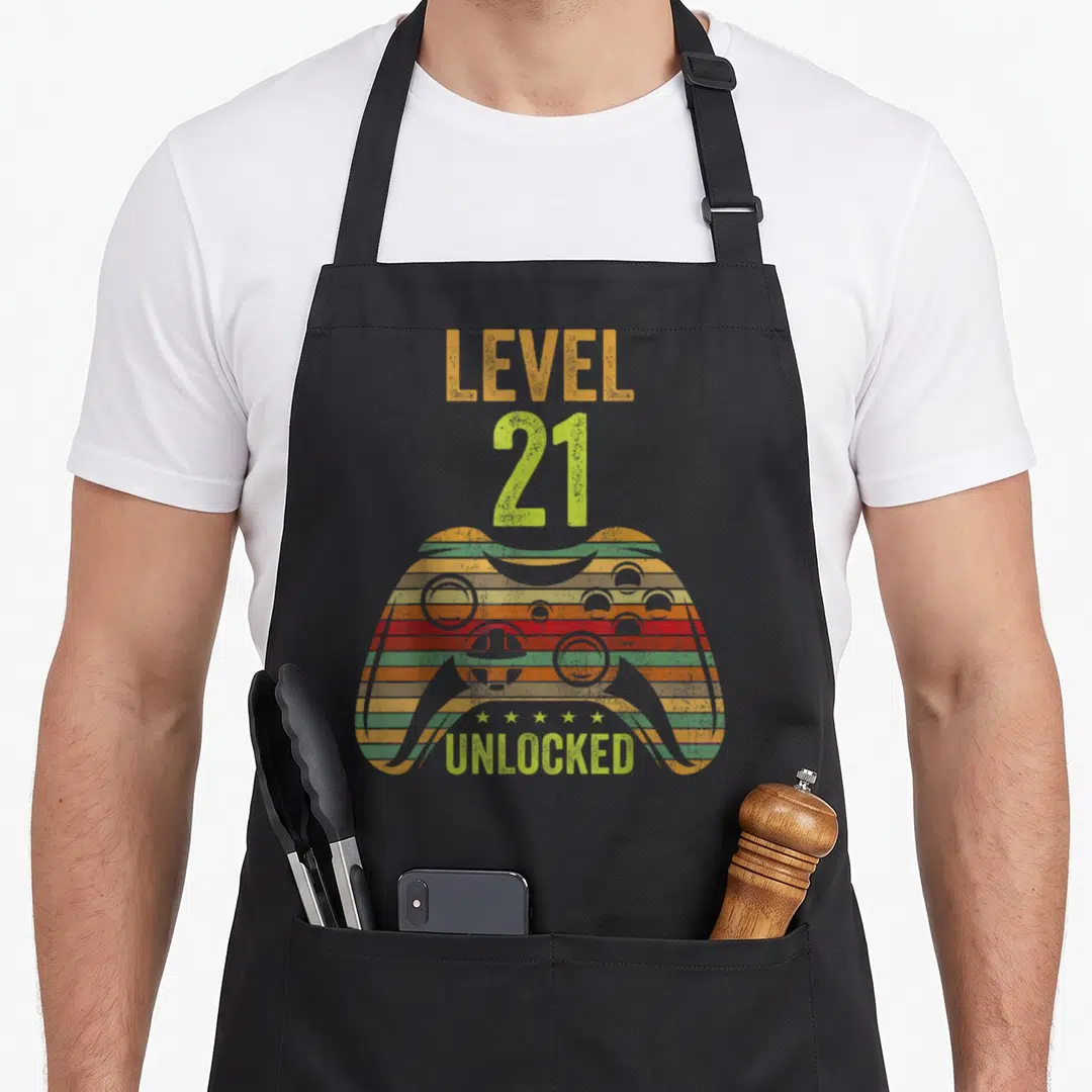 Cool level birthday apron - Striped game controller with LEVEL 21 UNLOCKED text and five stars in distressed orange red — shown on a black cotton canvas apron against a white background