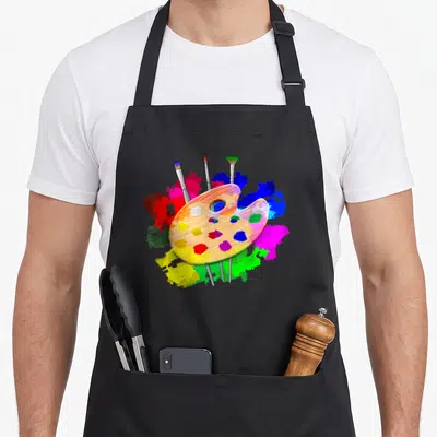 Artist paint apron - Cartoon tan palette with red blue yellow green pink blobs and three leaning brushes — shown on a black cotton canvas apron against a white background