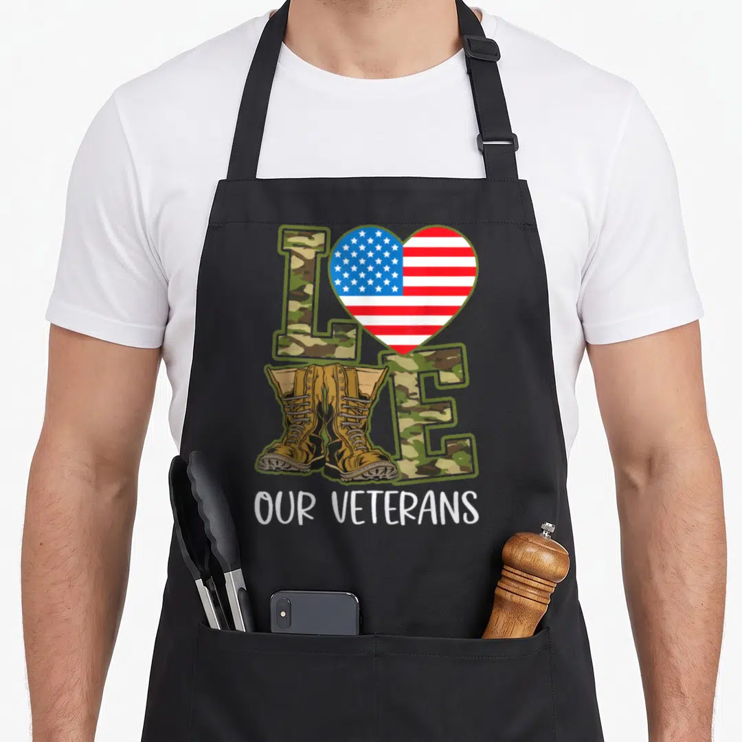 Our veteran military apron - LOVE text with camo letters, US flag heart, and tan boots above OUR VETERANS — shown on a black cotton canvas apron against a white background