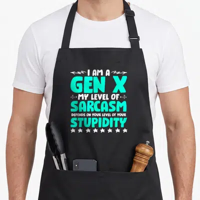 Level x apron - Stacked turquoise and white text reading I AM A GEN X with arrows and stars — shown on a black cotton canvas apron against a white background