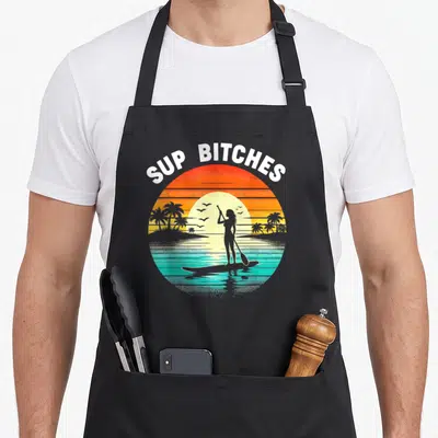 Paddleboard boarding apron - Retro sunset circle with woman on paddleboard and arched SUP BITCHES text in white — shown on a black cotton canvas apron against a white background