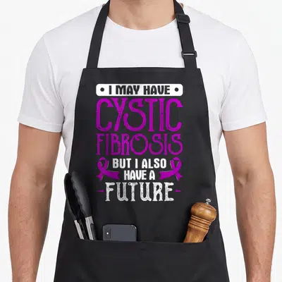 Fibrosis awareness ribbon apron - Purple and white stacked text reading I may have cystic fibrosis but I also have — shown on a black cotton canvas apron against a white background