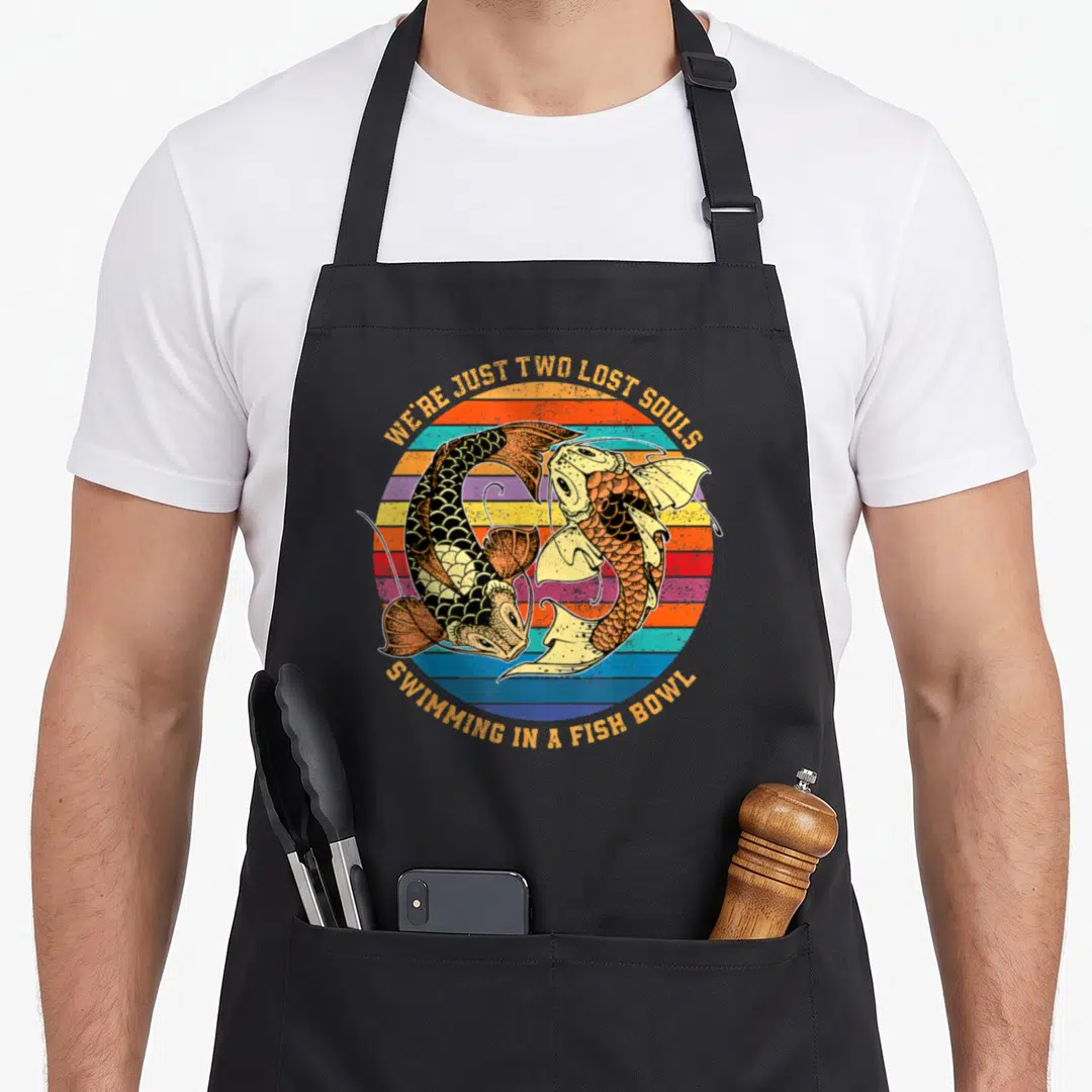 Soul fish apron - Two koi fish circling a striped retro sun with arched text WE'RE JUST TWO LOST SOULS — shown on a black cotton canvas apron against a white background
