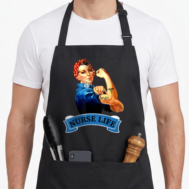 Nurse rosie the riveter apron - Retro woman flexing arm with Nurse tattoo and blue NURSE LIFE banner — shown on a black cotton canvas apron against a white background