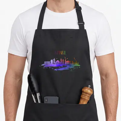 Souvenir turkey apron - ISTANBUL text above purple blue green red yellow watercolor skyline silhouette — shown on a black cotton canvas apron against a white background