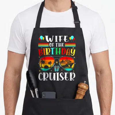 Sunglass wife apron - Wife of the Birthday Cruiser text with retro sunglasses showing a ship and palm trees — shown on a black cotton canvas apron against a white background