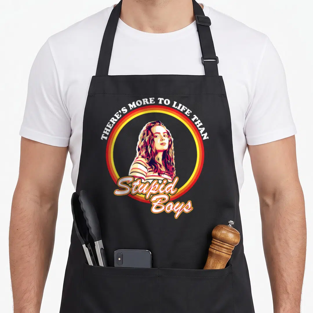 Max stupid aprons - Retro woman portrait in red yellow circle with THERE'S MORE TO LIFE THAN Stupid Boys text — shown on a black cotton canvas apron against a white background