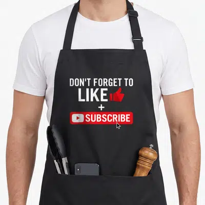 Media social apron - White text DON'T FORGET TO LIKE + SUBSCRIBE with red thumbs up and subscribe button graphic — shown on a black cotton canvas apron against a white background