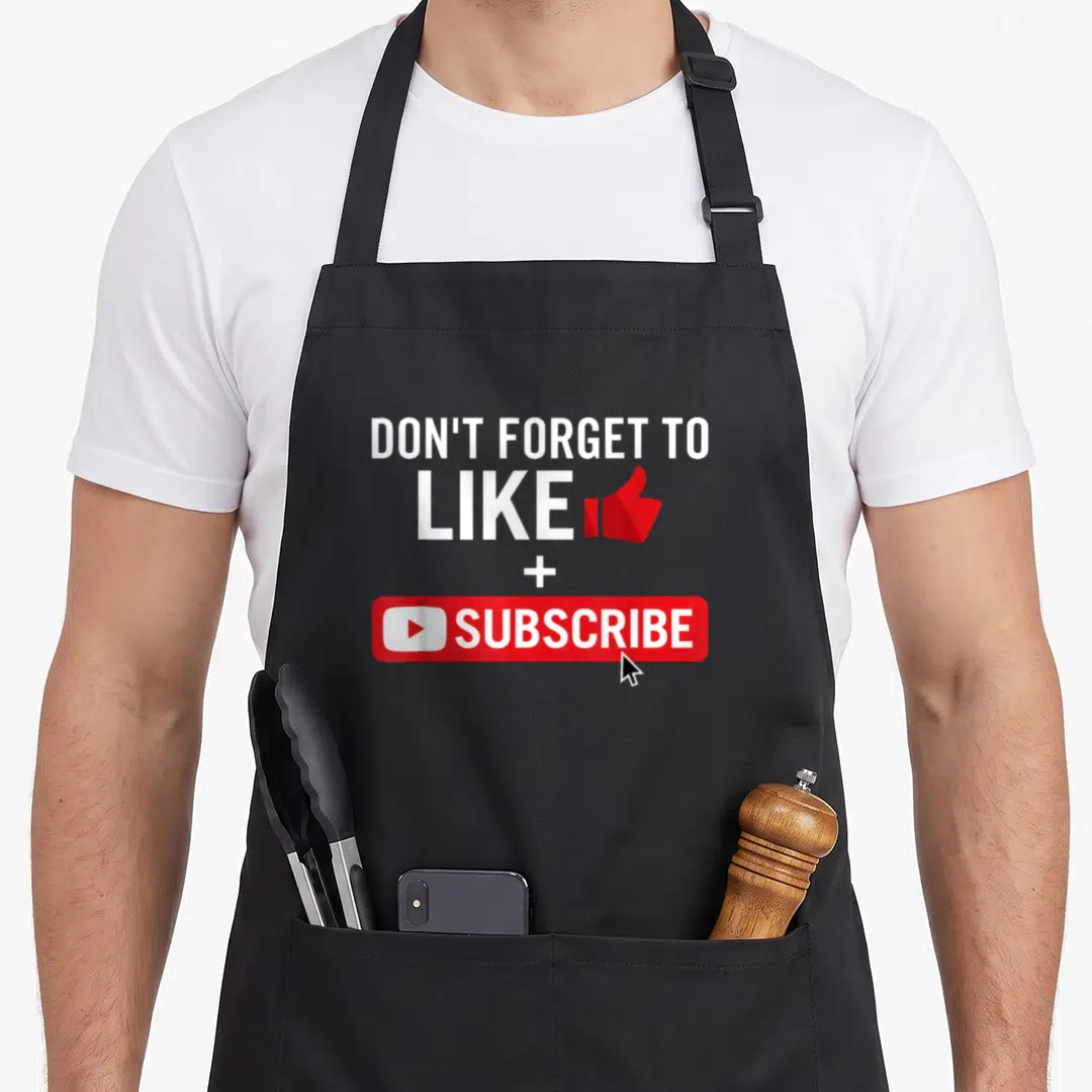 Media social apron - White text DON'T FORGET TO LIKE + SUBSCRIBE with red thumbs up and subscribe button graphic — shown on a black cotton canvas apron against a white background