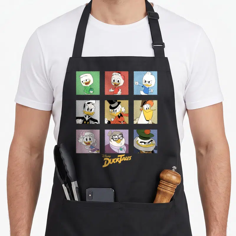 Ducktale box apron - Nine cartoon ducks in colored grid squares with centered logo text — shown on a black cotton canvas apron against a white background