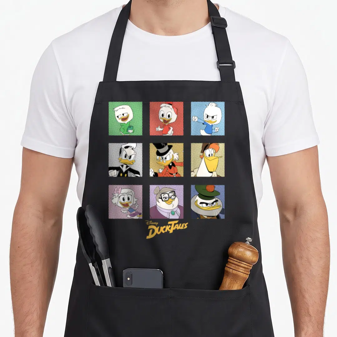Ducktale box apron - Nine cartoon ducks in colored grid squares with centered logo text — shown on a black cotton canvas apron against a white background