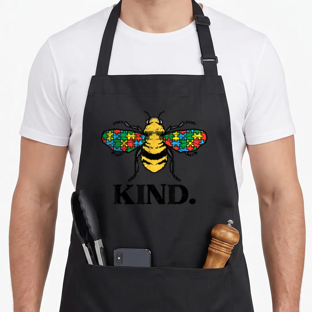 Puzzle bee apron - Cartoon bee with multicolored puzzle wings and large black KIND. text below — shown on a black cotton canvas apron against a white background
