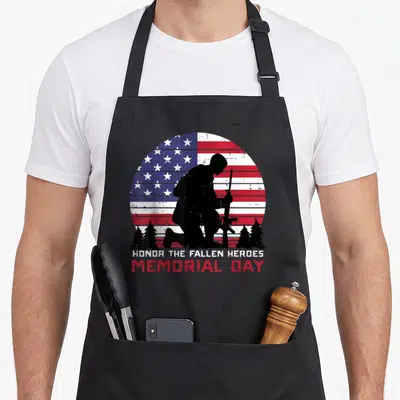 Honor us aprons - Kneeling soldier silhouette on distressed US flag with pine trees and memorial day text — shown on a black cotton canvas apron against a white background