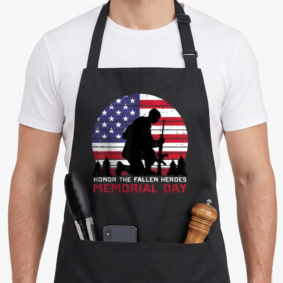 Honor us aprons - Kneeling soldier silhouette on distressed US flag with pine trees and memorial day text — shown on a black cotton canvas apron against a white background