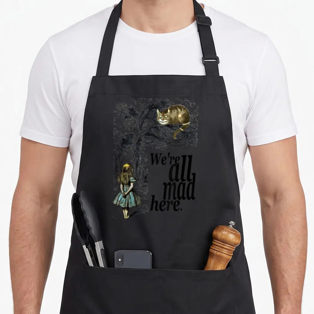 Funny mad alice wonderland apron - Cheshire cat perched on branch above girl facing away with text we're all mad here — shown on a black cotton canvas apron against a white background