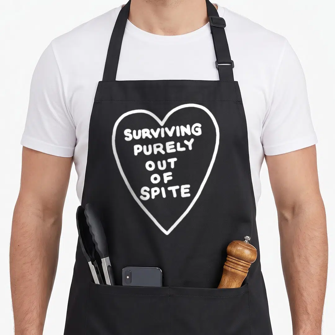 Surviving survivalist apron - White heart outline containing stacked text reading Surviving Purely Out of Spite — shown on a black cotton canvas apron against a white background