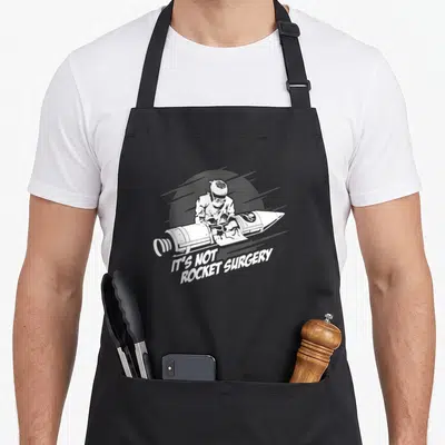 Rocket surgeon apron - Surgeon operating on a rocket with text IT'S NOT, ROCKET SURGERY in white line art — shown on a black cotton canvas apron against a white background